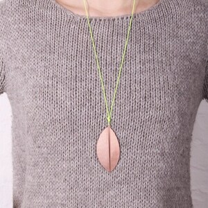 May include: A copper-colored leaf pendant hangs from a neon green cord necklace. The pendant is a stylized leaf shape with a vertical crease down the center. The necklace is worn over a gray knit sweater.