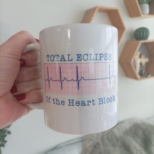 May include: White ceramic mug with a blue and pink design. The mug features a heart rate monitor graphic and the text "TOTAL ECLIPSE Of the Heart Block".