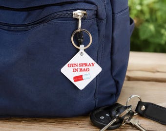 Puede incluir: Una etiqueta de llavero blanca en forma de diamante sujeta a la cremallera de un bolso azul marino. La etiqueta presenta el texto rojo "GTN SPRAY IN BAG" sobre un gráfico de un spray rojo y blanco. Un anillo de metal plateado conecta la etiqueta.