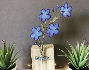 Puede incluir: Un jarrón de pared de madera con un tubo de ensayo de vidrio transparente para sostener un pequeño ramo de flores de papel azul. El jarrón está montado en un bloque de madera con soportes metálicos.