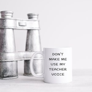 Puede incluir: Taza de caf&eacute; de cer&aacute;mica blanca con texto negro que dice "No me hagas usar mi voz de maestro". La taza est&aacute; sobre una superficie blanca junto a un par de binoculares plateados.