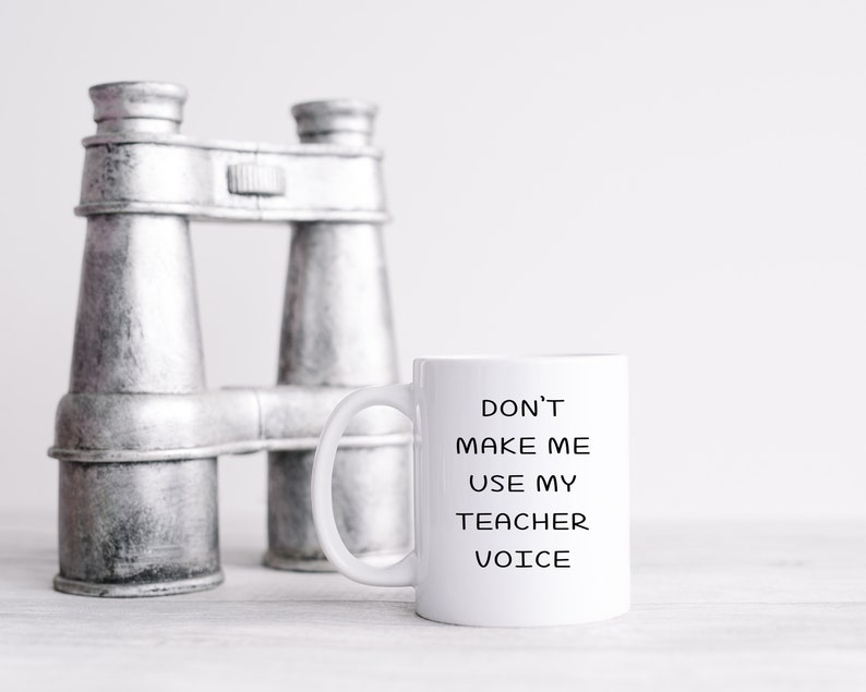 Puede incluir: Taza de caf&eacute; de cer&aacute;mica blanca con texto negro que dice "No me hagas usar mi voz de maestro". La taza est&aacute; sobre una superficie blanca junto a un par de binoculares plateados.