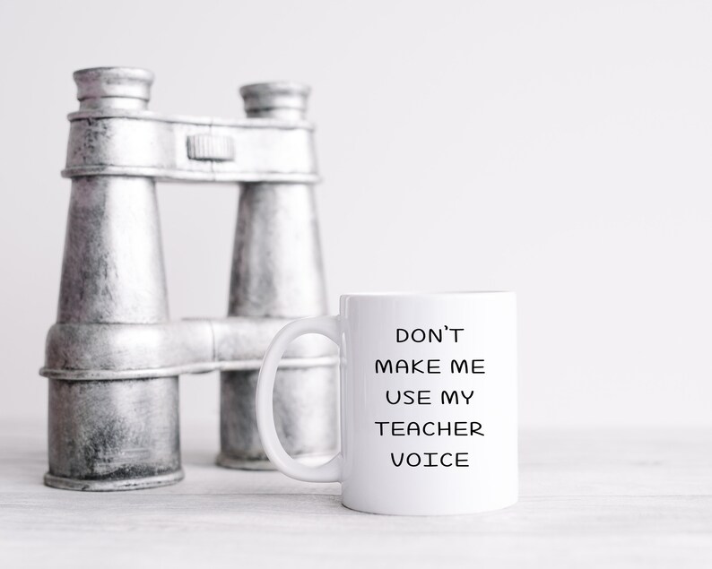 Puede incluir: Taza de caf&eacute; de cer&aacute;mica blanca con texto negro que dice "No me hagas usar mi voz de maestro". La taza est&aacute; sobre una superficie blanca junto a un par de binoculares plateados.