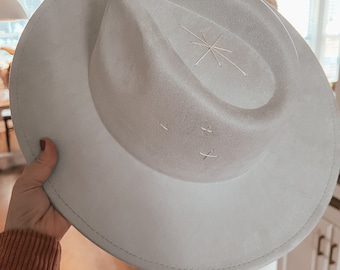 Pode incluir: Um chapéu fedora cinza-claro com aba larga e copa vincada. O chapéu apresenta detalhes bordados em branco, incluindo um design de estrela no topo da copa e dois pequenos padrões de ponto cruz na lateral da copa.