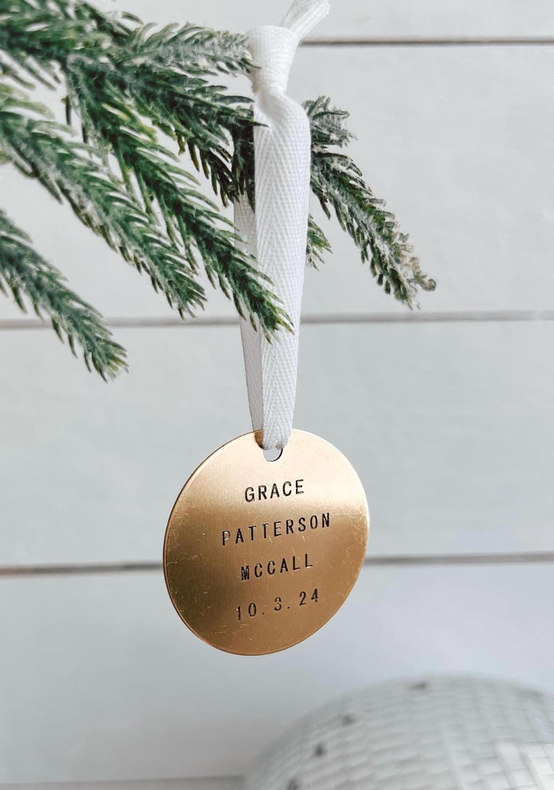 以下が含まれることがあります： 「GRACE PATTERSON MCCALL 10.3.24」と刻まれた丸い金色のオーナメント。白いリボンで吊り下げられています。