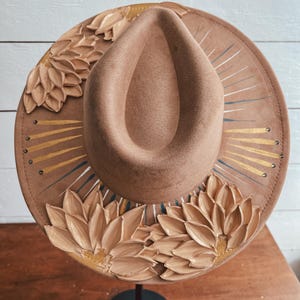 May include: A brown felt hat with a wide brim, decorated with 3D sculpted flowers and gold accents. The hat is on a black stand.