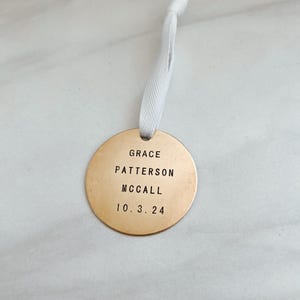 以下が含まれることがあります： 白いリボンが付いた丸い金色のオーナメント。オーナメントには「GRACE PATTERSON MCCALL 10.3.24」という文字が刻まれています。