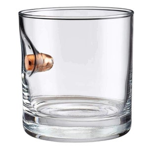 Puede incluir: Un vaso de vidrio transparente con una bala incrustada en el lateral. La bala es de color cobre y tiene una punta redondeada.