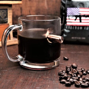 May include: A clear glass mug with a copper bullet embedded in the side. The mug is filled with dark coffee and is sitting on a wooden surface with coffee beans scattered around it.