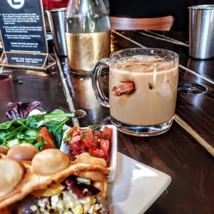 May include: A close-up of a glass mug with a wooden handle, filled with iced coffee. The mug is sitting on a wooden table next to a plate with a waffle, salsa, and salad.