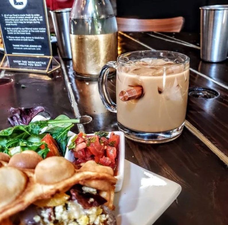 May include: A close-up of a glass mug with a wooden handle, filled with iced coffee. The mug is sitting on a wooden table next to a plate with a waffle, salsa, and salad.