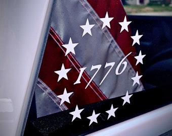 May include: A white decal of thirteen stars in a circle surrounding the year 1776, placed on a black surface with a reflection of an American flag in the background.