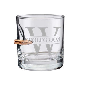 May include: A clear glass whiskey tumbler with a bullet embedded in the side. The tumbler is engraved with the letter "W" and the name "WOLFGAM".