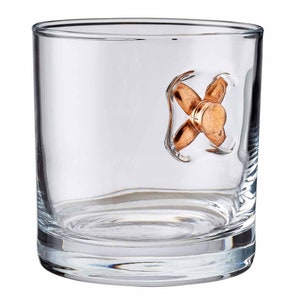 Puede incluir: Un vaso de vidrio transparente con una bala de cobre incrustada en el lateral. La bala est&aacute; colocada en un ligero &aacute;ngulo, con la punta mirando hacia el lado derecho del vaso.