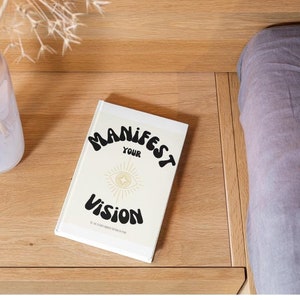 Może przedstawiać: Biała książka z tekstem "MANIFEST YOUR VISION" w czarnych, stylizowanych literach. Książka ma złotą ilustrację oka z gwiazdą w środku.