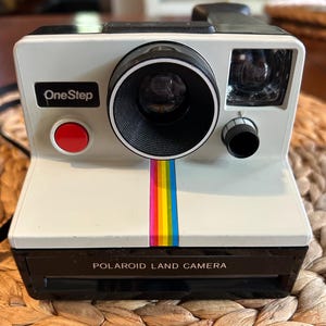 Op de afbeelding: Witte Polaroid OneStep Land Camera met een zwarte lens, flitser en handvat. Een verticale regenboogstreep loopt over de voorkant. De camera heeft een rode knop en de tekst "OneStep" en "POLAROID LAND CAMERA".