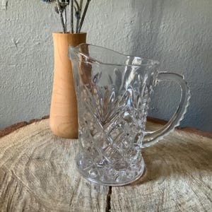 May include: A clear glass pitcher with a diamond pattern and a handle. Beside it is a small wooden vase holding blue thistle-like flowers. The items are on a wooden surface.