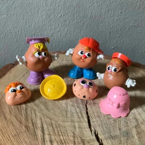 May include: A set of six vintage Mr. Potato Head toys, including a purple and pink potato head wearing a graduation cap, a red and blue potato head wearing a baseball cap, a brown potato head wearing glasses, a yellow hat, a pink hat, and a brown potato head with a pink nose and purple eyes.