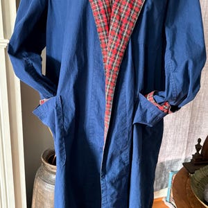 May include: A long, navy blue robe with a red, black, and white plaid collar and cuffs. The robe has two front pockets and is displayed on a mannequin. The robe is made of a soft fabric.