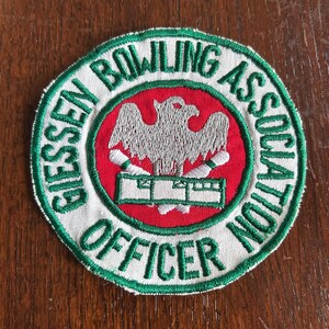 May include: Circular patch with the text "GIESSEN BOWLING ASSOCIATION OFFICER" in green on a white background. The center features a red circle with a gray eagle and bowling pins.