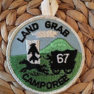 May include: A round, green and blue embroidered patch with white stitching. The patch features a black horse-drawn wagon with the number "67" on the side. The text "Land Grab Camporee" is embroidered in black on the patch.