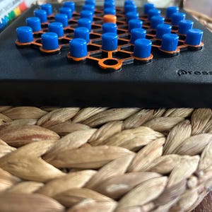 May include: A black board game with blue and orange pegs arranged in a grid. One orange peg is in the center. The game is set on a woven placemat.