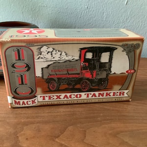 Op de afbeelding: Een vintage 1910 Mack Texaco Tanker spaarpot met sleutel, van gegoten metaal. De doos is bruin met rode en witte accenten. De vrachtwagen is afgebeeld in zwart-wit met een rood Texaco-logo.