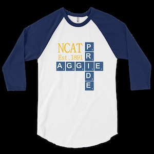 May include: White and navy blue raglan baseball shirt with the text "NCAT Aggie Pride" in gold and blue. The shirt has three-quarter length sleeves and a curved hemline. The text includes the founding year, 1891.