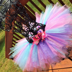May include: A colorful tutu skirt in shades of pink, purple, and blue, displayed on a black metal dress form. The skirt features layers of tulle and a decorative bow with a character design.
