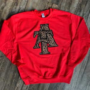May include: A bright red sweatshirt with a crew neck. The front features a large, stylized "ATA" design in a leopard print pattern with black trim. The sweatshirt is made of a soft material.