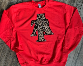 Könnte beinhalten: Ein leuchtend rotes Sweatshirt mit Rundhalsausschnitt. Auf der Vorderseite befindet sich ein großes, stilisiertes "ATA"-Design im Leopardenmuster mit schwarzem Rand. Das Sweatshirt ist aus weichem Material.