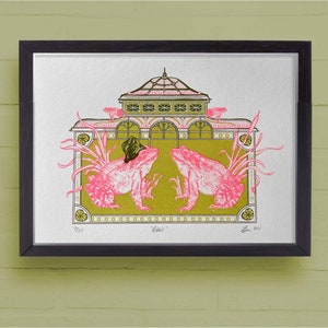 May include: Framed art print depicting two pink frogs wearing hats, facing each other, set against a building with a green and pink design. The artwork has a vintage style, with the word "Odd" and the artist's signature visible.