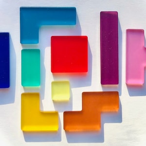 May include: A collection of colorful, translucent geometric shapes in various forms, including squares, rectangles, and L-shapes. The pieces are in shades of blue, green, red, yellow, orange, purple, and pink, arranged on a white surface.