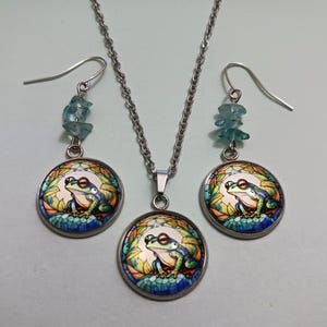 May include: A silver-toned necklace and earring set featuring a frog design. The pendant and earrings have a round, colorful frog illustration. The earrings have blue gemstone accents. The frog is green, blue, and white.