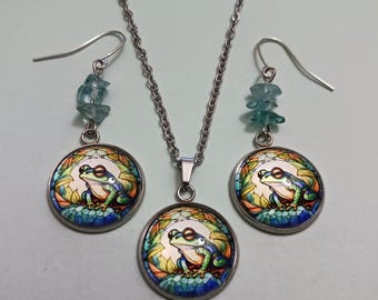 May include: A silver necklace and earring set with a frog design. The pendant and earrings feature a round, colourful frog illustration. The earrings have blue gemstone accents. The frog is green, blue, and white.