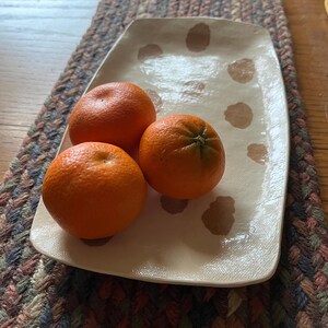 May include: Three bright orange tangerines rest on a rectangular, off-white ceramic plate with brown polka dots. The plate sits on a braided, multi-colored table runner, creating a rustic, natural aesthetic.