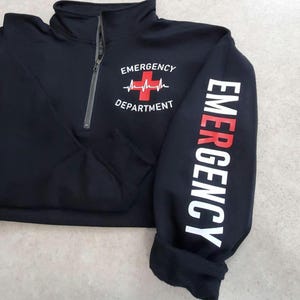 May include: Black quarter-zip sweatshirt with the words "EMERGENCY DEPARTMENT" printed on the front and sleeve. The design includes a red cross and heartbeat graphic. The sweatshirt has a gray zipper.