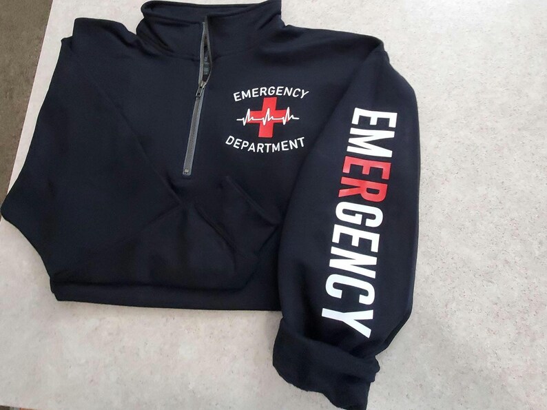 May include: Black quarter-zip sweatshirt with the words "EMERGENCY DEPARTMENT" printed on the front and sleeve. The design includes a red cross and heartbeat graphic. The sweatshirt has a gray zipper.