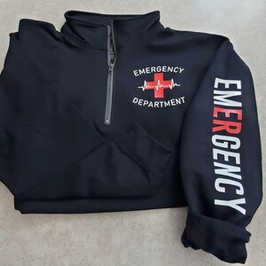 May include: Black quarter-zip sweatshirt with "EMERGENCY DEPARTMENT" printed on the front and "EMERGENCY" down the sleeve. The design includes a red cross and heartbeat graphic. The zipper is silver.