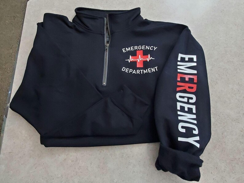 May include: Black quarter-zip sweatshirt with "EMERGENCY DEPARTMENT" printed on the front and "EMERGENCY" down the sleeve. The design includes a red cross and heartbeat graphic. The zipper is silver.