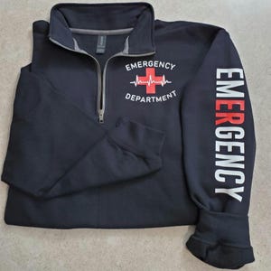 May include: Navy blue quarter-zip sweatshirt with the words "EMERGENCY DEPARTMENT" and a red cross graphic on the front. The right sleeve has the word "EMERGENCY" printed vertically in white and red. The sweatshirt has a gray collar.