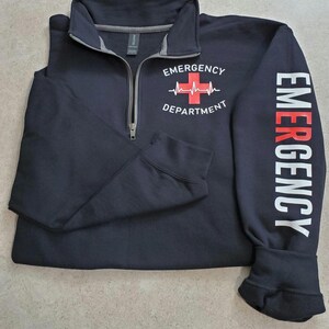 May include: Navy blue quarter-zip sweatshirt with "EMERGENCY DEPARTMENT" printed on the chest, featuring a red cross and heartbeat graphic. The right sleeve displays the word "EMERGENCY" in white. A practical and comfortable garment.