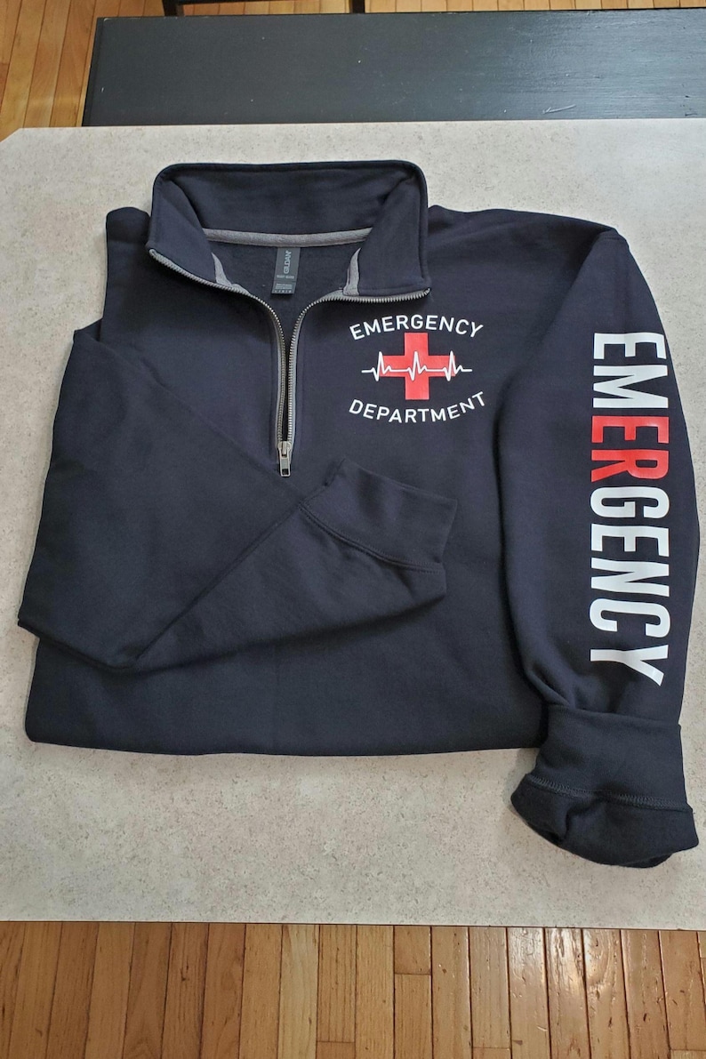 May include: Navy blue quarter-zip sweatshirt with "EMERGENCY DEPARTMENT" printed on the chest, featuring a red cross and heartbeat graphic. The right sleeve displays the word "EMERGENCY" in white. A practical and comfortable garment.