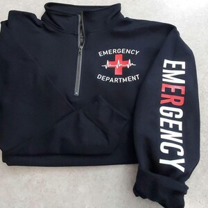 May include: Black quarter-zip sweatshirt with "EMERGENCY DEPARTMENT" printed on the front and "EMERGENCY" down the sleeve. The design includes a red cross and heartbeat graphic. The sweatshirt has a high collar and a silver zipper.