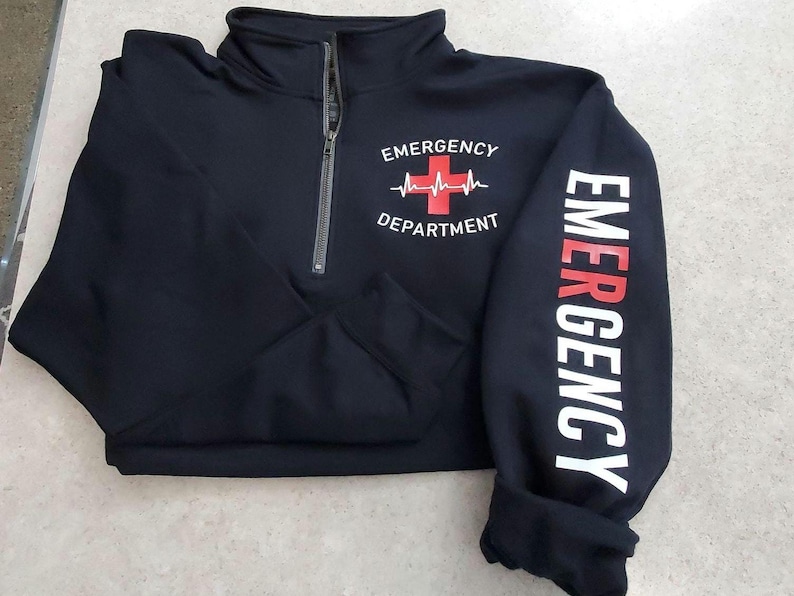 May include: Black quarter-zip sweatshirt with "EMERGENCY DEPARTMENT" printed on the front and "EMERGENCY" down the sleeve. The design includes a red cross and heartbeat graphic. The sweatshirt has a high collar and a silver zipper.