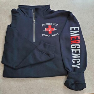 May include: Navy blue quarter-zip sweatshirt. The front features the text "EMERGENCY DEPARTMENT" with a red cross and heartbeat graphic. The right sleeve displays the word "EMERGENCY" in white and red vertical lettering. The sweatshirt has a silver zipper.