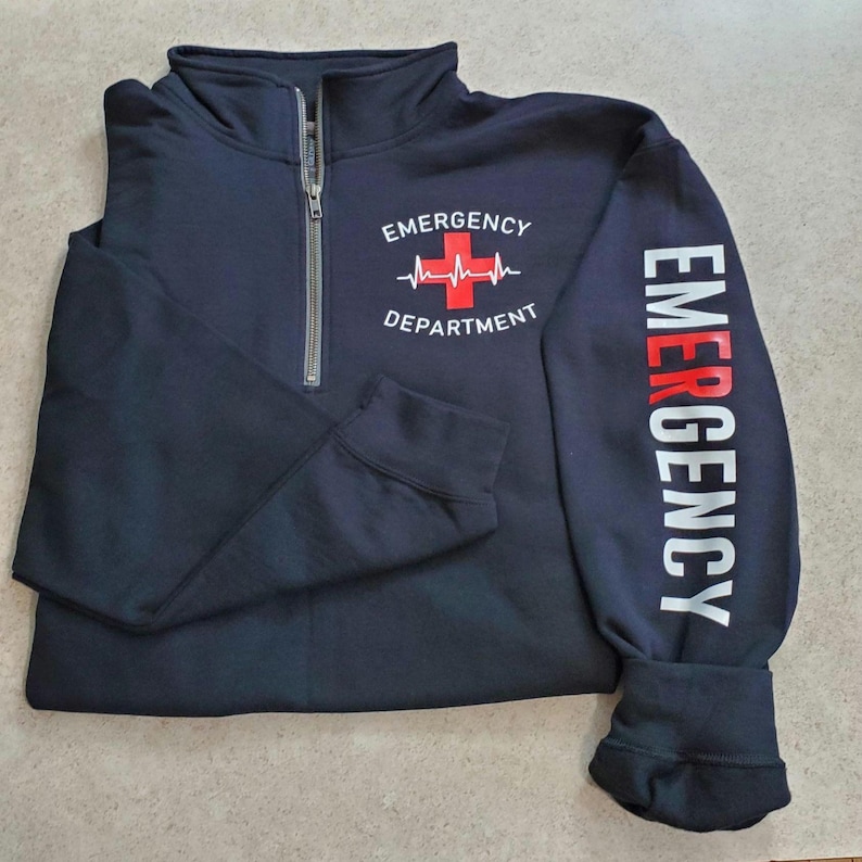 May include: Navy blue quarter-zip sweatshirt. The front features the text "EMERGENCY DEPARTMENT" with a red cross and heartbeat graphic. The right sleeve displays the word "EMERGENCY" in white and red vertical lettering. The sweatshirt has a silver zipper.