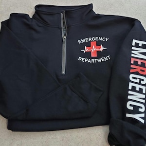 May include: Black quarter-zip sweatshirt with the words "Emergency Department" and a red cross with a heartbeat graphic on the front. The word "EMERGENCY" is printed vertically in white on the left sleeve.