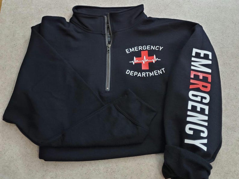 May include: Black quarter-zip sweatshirt with the words "Emergency Department" and a red cross with a heartbeat graphic on the front. The word "EMERGENCY" is printed vertically in white on the left sleeve.