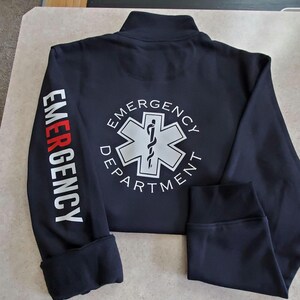 May include: Black zippered jacket with long sleeves and white text on the sleeves and back. The text on the sleeves reads "EMERGENCY". The text on the back reads "EMERGENCY DEPARTMENT" with a white star of life graphic.